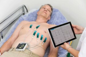 Doctor checks ECG results on tablet, male patient lies nearby